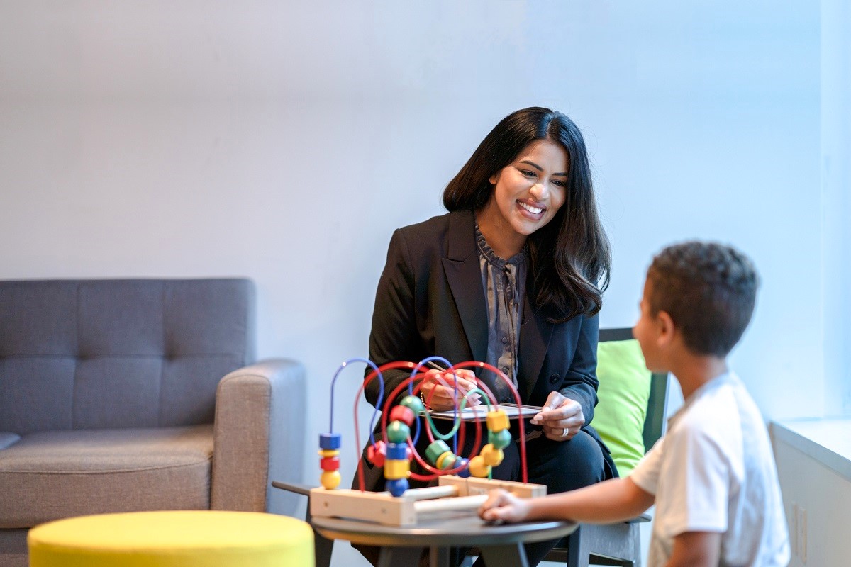 A female ABA therapist sits in chair beside young patient as they play together with a bead maze