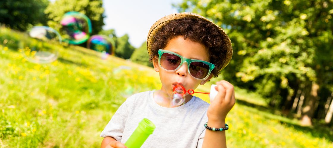 Child in field during summer blowing bubbles while wearing sunglasses and hat